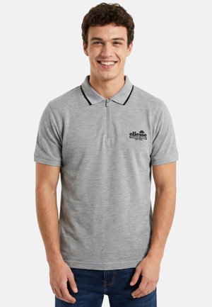 Jeune homme souriant portant un polo gris clair à manches courtes avec une fermeture éclair noire et un logo sur le côté gauche de la poitrine.