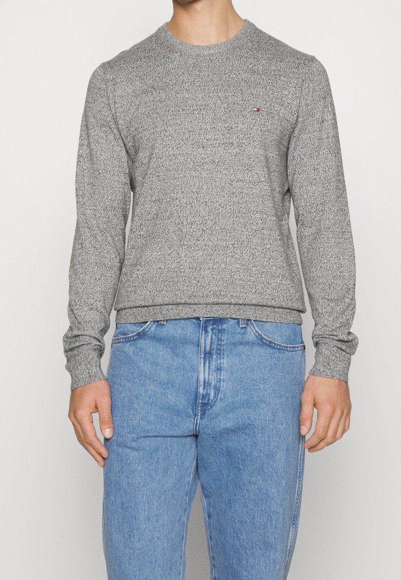 Homme portant un pull gris à manches longues et col rond, ainsi qu'un jean bleu clair, debout devant un fond blanc uni.
