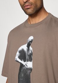 Pruun puuvillane T-särk, mille esiküljel on suur must-valge graafiline trükk figuurist. Lühikesed varrukad, ümar kaelus, mugav lõige.