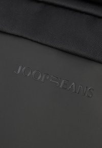 Textil negro con el logo en relieve "JOOP JEANS". El material es suave, con un ligero brillo y una tela texturizada superpuesta.