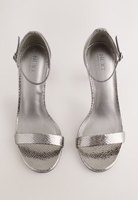 Srebrne sandały na wysokim obcasie z teksturą wężowego wzoru, odkrytymi palcami i paskiem wokół kostki. Gładkie metaliczne wykończenie na podeszwie i pasku.
