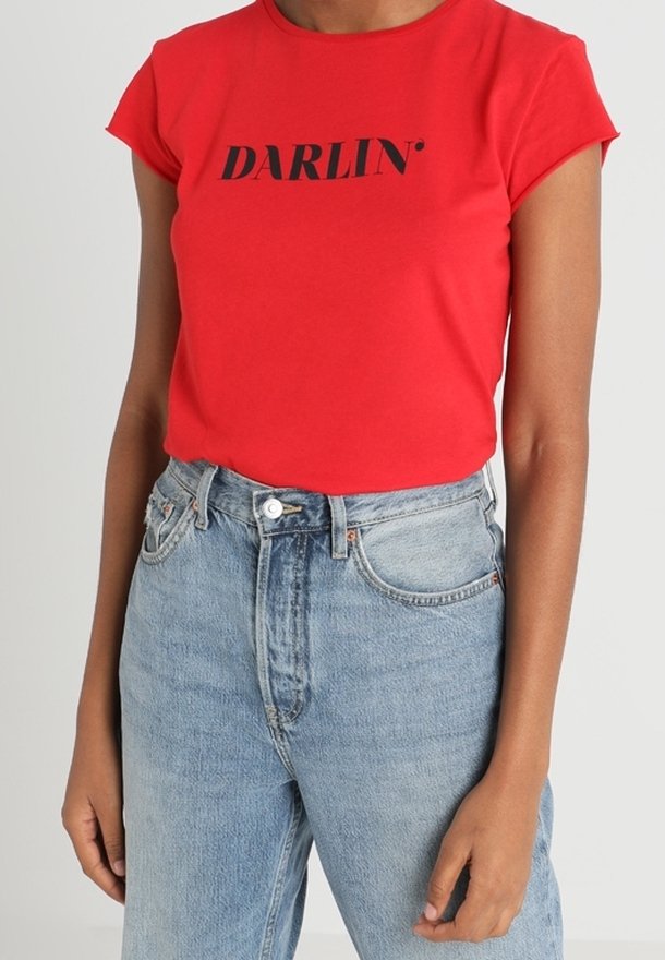 Rood T-shirt met korte mouwen en de tekst "DARLIN'" in het zwart, gecombineerd met een lichtblauwe high-waisted spijkerbroek bij een persoon.