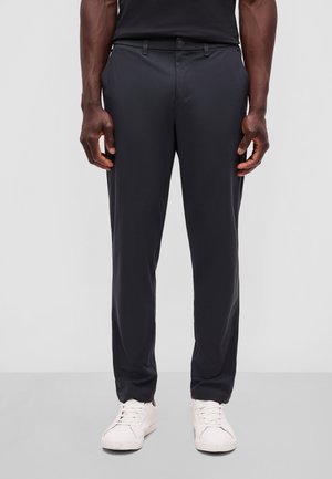 Mand iført sorte slim-fit bukser og hvide sneakers, stående mod en ensfarvet lys baggrund.