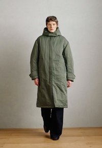 Cappotto imbottito lungo, verde oliva con colletto alto e cappuccio. Presenta maniche larghe e una silhouette dritta. Texture liscia e opaca.