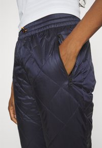 Marina quiltade byxor med elastisk midja och justerbar snodd, sidoficka med dragkedja och slät, glänsande tygtextur.