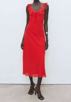 Femme portant une robe midi rouge sans manches avec un col volanté et des sandales noires à talons, debout sur un sol clair contre un mur blanc.