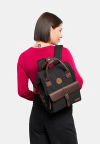 Sac à dos en toile noire avec des accents en cuir brun, une patte de fermeture à l'avant sécurisée par une boucle en métal, et un patch logo rond. Comprend des poignées doubles et des poches latérales.