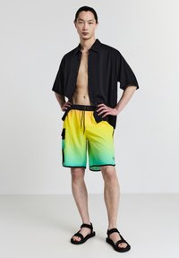 Schwarzes kurzärmliges Hemd, unsteif sitzen. Neon-gelbe bis grüne Verlauf-Badeshorts mit schwarzen Akzenten und Seitentaschen. Schwarze Sandalen.