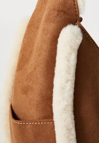 Gros plan sur un tissu en suede marron avec une bordure en shearling blanche et du fil visible sur le bord de la poche d'un vêtement.