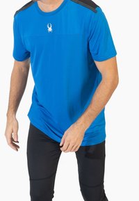 Camisa atlética azul con un cuerpo sólido y detalles negros en los hombros; presenta un pequeño logotipo de araña blanca en el pecho. Textura suave.