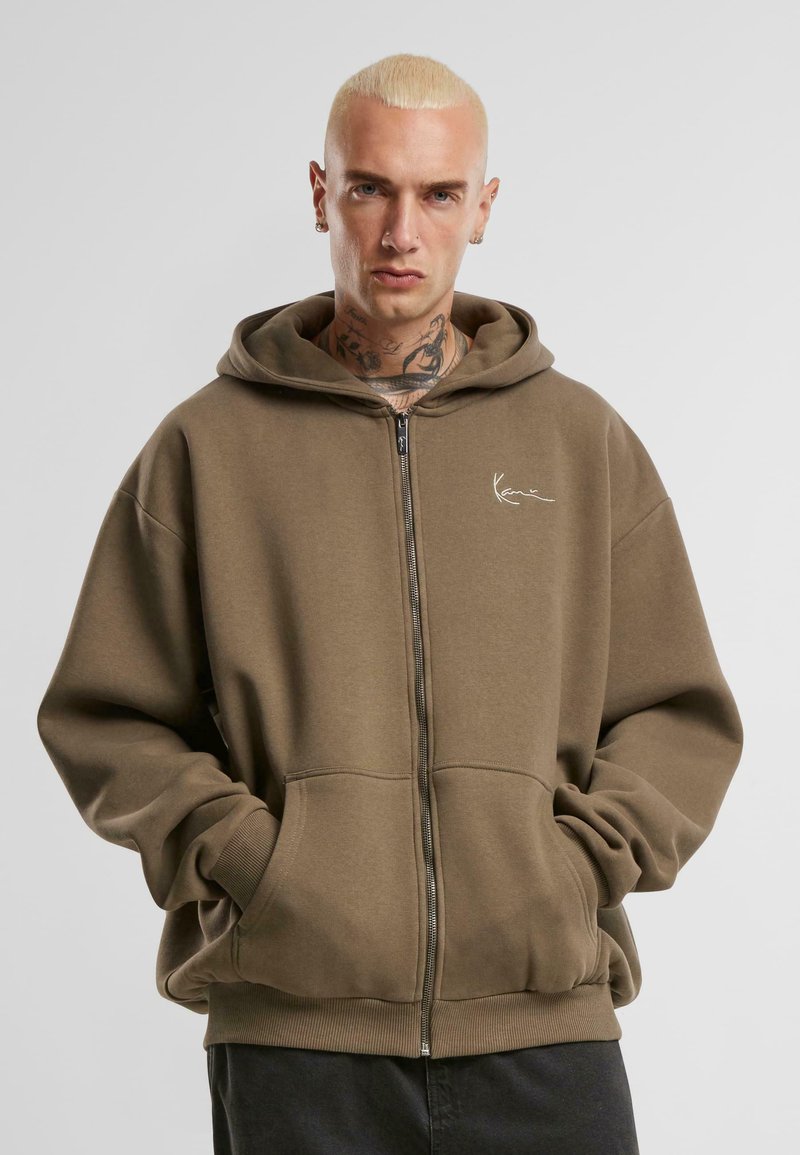 Sweatshirt zippé à capuche en vert olive, fabriqué en tissu doux, avec une poche avant, des poignets côtelés et un logo brodé sur la poitrine.