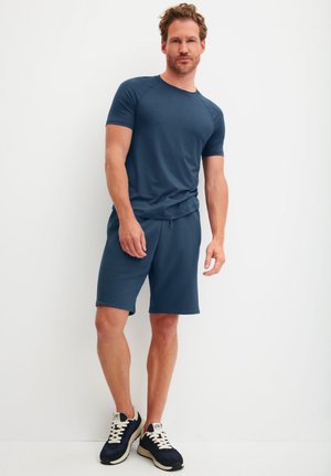 Mann steht in marineblauem Sportshirt und Shorts mit schwarzen und weißen Turnschuhen vor einer schlichten weißen Wand.