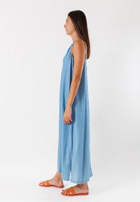 Abito lungo in denim blu senza maniche, con spacchi laterali, una vestibilità rilassata e spalline sottili. Abbinato a sandali piatti arancioni.