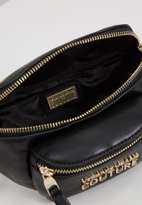 Černá kožená taštička se zlatými zipy, přední kapsou a zlatým logem "Versace Jeans Couture" na přední straně a vnitřním štítku.