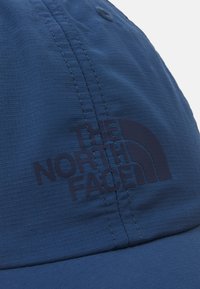 Boné azul feito de um tecido liso e texturado com um logótipo escuro da "The North Face" e um detalhe de design semicircular.