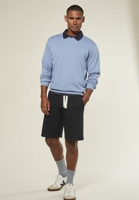 Mann trägt einen hellblauen Langarmoberteil mit navyfarbenem Kragen, schwarze Shorts mit Kordelzug, graue Socken und weiße Turnschuhe, während er vor einem schlichten Hintergrund steht.