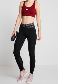 Tenue de sport Nike comprenant un soutien-gorge de sport bordeaux, un legging noir avec une ceinture de marque, et des baskets roses. Matériaux lisses et extensibles.