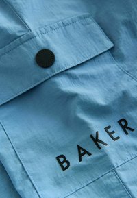 Jasnoniebieska tkanina z kieszenią na klapę zabezpieczoną czarnym guzikiem z wytłoczonym napisem "Baker by Ted Baker" oraz czarnym napisem "BAKER" na kieszeni.