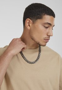 Zilveren metalen ketting met grote schakels, gedragen over een beige katoenen t-shirt. Donker haar, gezien vanaf de zijkant. Eenvoudige achtergrond.