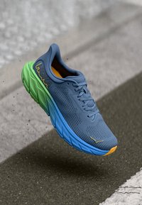 Chaussure de sport bleue et verte avec une tige en maille respirante, un branding jaune distinctif et une semelle texturée, dotée d'un design rembourré.