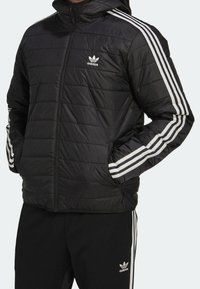 Zwarte pufferjas met capuchon, voorzien van horizontale stiknaden, witte drie-strepen accenten op de mouwen en ritszakken aan de voorkant.