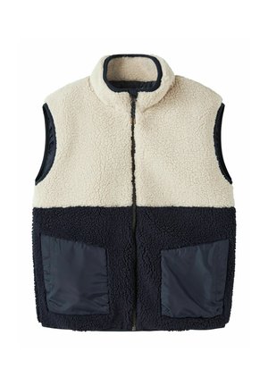 Veste sans manches en polaire sherpa avec partie supérieure crème et partie inférieure noire, équipée d'une fermeture éclair à l'avant et de deux poches en tissu noires.