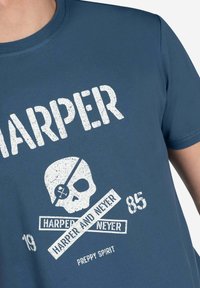 Camiseta de algodón azul con un gran gráfico blanco que presenta una calavera y tibias cruzadas, el texto "HARPER AND NEYER" y detalles desgastados. Manga corta.