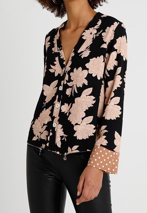 Vrouw draagt een zwarte blouse met knopen, grote beige bloemmotieven en beige polkadot manchetten, gecombineerd met een zwarte leren broek.