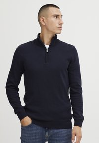 Navy Strickpullover mit einem Reißverschluss am Stehkragen, langen Ärmeln und gerippten Bündchen. Getragen über einem weißen Hemd und kombiniert mit blauen Jeans.