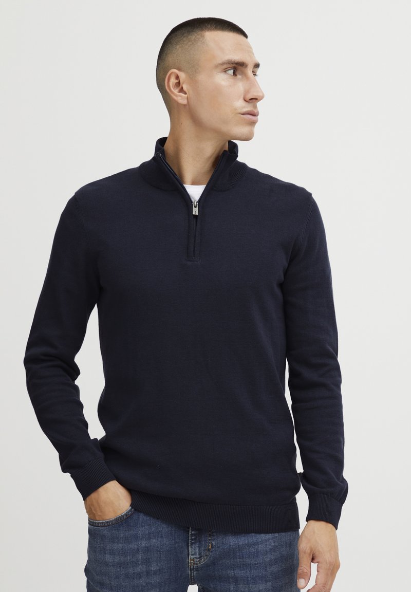 Navy Strickpullover mit einem Reißverschluss am Stehkragen, langen Ärmeln und gerippten Bündchen. Getragen über einem weißen Hemd und kombiniert mit blauen Jeans.