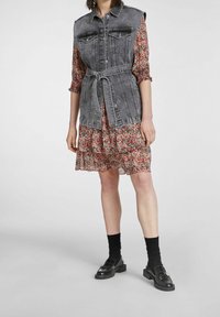 Veste en jean grise, avec fermeture à boutons et poches avant, superposée sur une robe à fleurs volantes rouge et verte, associée à des mocassins noirs et des chaussettes.