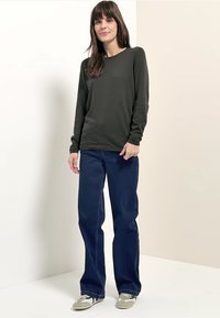 Vrouw die een donkergroene top met lange mouwen, wijde donkerblauwe jeans en witte sneakers met groene strepen draagt, staand tegen een neutrale achtergrond.