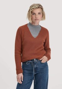 Rustfarbenes gestricktes Sweatshirt mit tiefem V-Ausschnitt, gerippter Textur und langen Ärmeln, kombiniert mit einem grauen Rollkragenpullover und blauen Jeans.