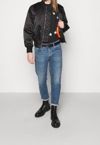 Černá saténová bomber bunda s žebrovanými manžetami, černý svetr s hvězdným vzorem, světle modré džíny a černé šněrovací boty.