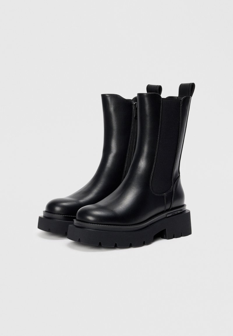 Stivali alla caviglia in pelle nera con suola piatta robusta, pannelli laterali elastici e linguette per una facile calzata. Texture liscia con dettagli di cucitura minimi.