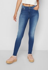 Jeans ajustados en denim azul con efecto desgastado. Cuenta con cinco bolsillos, pasadores para cinturón y cierre con cremallera. Llevados con zapatos de plataforma de color claro.