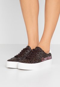 Brązowe sneakersy w wzory z białą gumową podeszwą, z brązowymi sznurowadłami i czerwonym logo "Calvin Klein" po boku. Teksturowany materiał wierzchni.