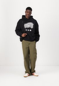 Sweatshirt preto com design gráfico, calças cargo verde oliva e sapatilhas creme com riscas vermelhas; apresenta uma alça e um corte descontraído.