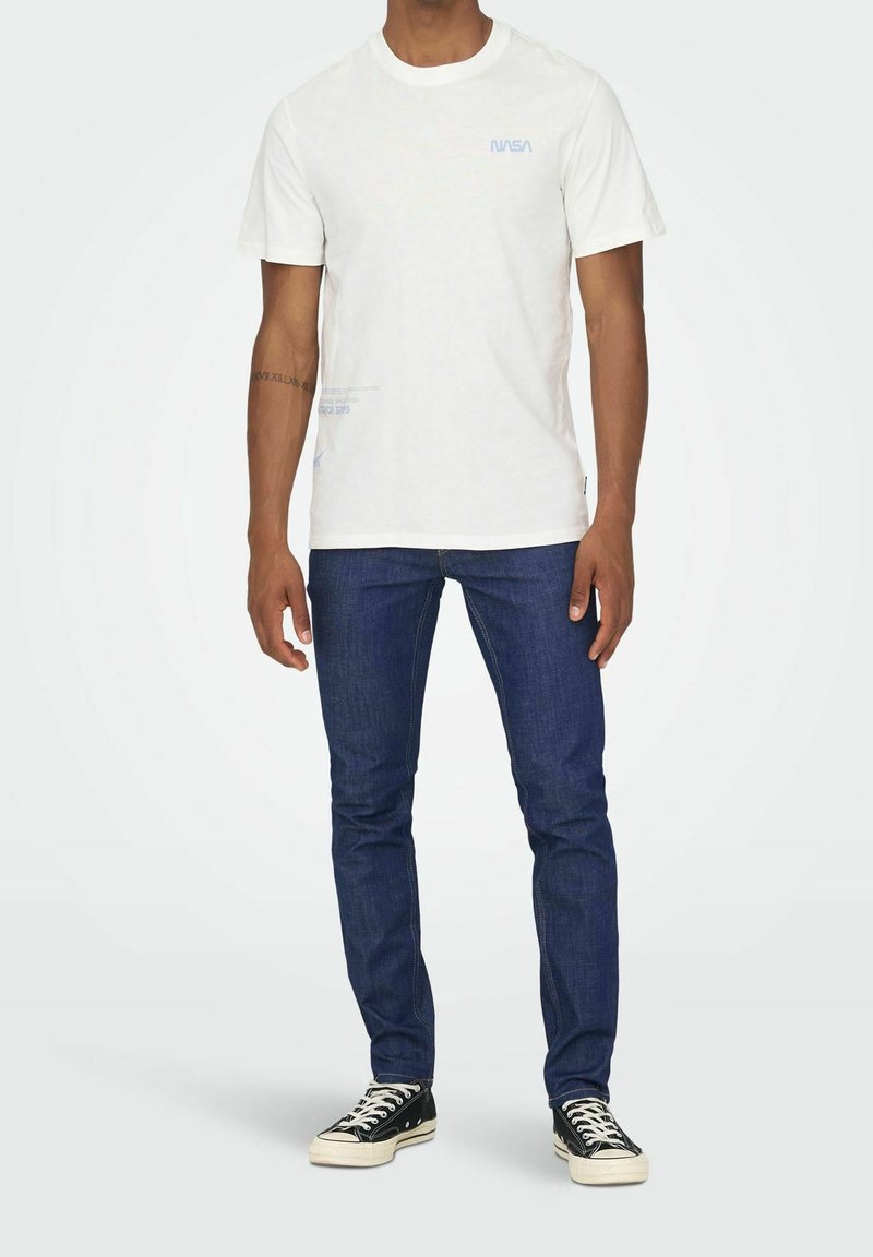 Homme portant un t-shirt blanc avec le logo NASA, jean slim bleu foncé et baskets noires, debout devant un fond blanc.