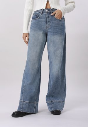 Person iført højtaljede, lyseblå viddebukser med syninger ved kanterne, sorte sko og en cropped hvid ribstrikket sweater med lynlås.