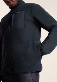 Dark green fleece jacket with a high collar, zip closure, and a contrasting black chest pocket; features side pockets and textured fabric.