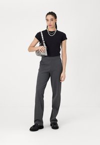 Chemise noire à manches courtes associée à un pantalon taille haute gris foncé. Le mannequin tient un petit sac métallique ; porte un collier de perles et des chaussures noires.