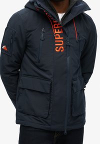 Homme portant une veste à capuche bleu marine foncé avec plusieurs poches zippées et un grand texte vertical orange "SUPER" à l'avant.