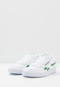 Bele snežne kože na belih čevljih z zelenimi poudarki, perforirano prstno škatlo, ravne vezalke in teksturiran gumjast podplat. Vsebuje Reebok logo na strani.