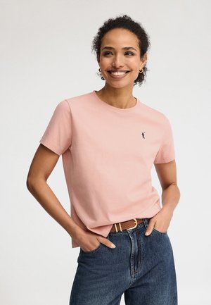 Uśmiechnięta kobieta o kręconych włosach, ubrana w różowy t-shirt z małym logo, niebieskie dżinsy i brązowy pasek, z rękami w kieszeniach na białym tle.