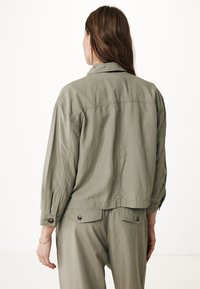 Giacca verde oliva chiaro realizzata in tessuto morbido, con una vestibilità comoda, polsini con bottoni e una spallina sul retro per dare struttura. Design minimalista senza motivi.