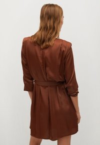 Brązowa sukienka z satyny, z paskiem w talii, trzema ćwierć rękawami i luźnym krojem. Materiał ma gładką, błyszczącą teksturę.