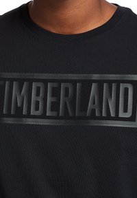 Camiseta negra con un destacado logo en gris de "TIMBERLAND" en el pecho, con una textura suave y mate y un cuello redondo.