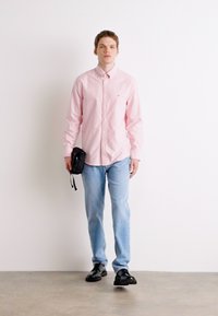 Lys rosa skjorte med knapp, lyseblå jeans, svarte sko og en liten svart veske. Står mot en hvit bakgrunn, med ansiktet fremover.