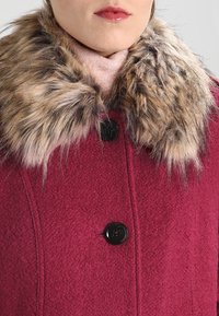 Burgundin villatakki, jossa on tekoturkis kaulus. Varustettu mustilla napilla ja teksturoidulla kankaalla. Lähikuva, joka esittelee kauluksen yksityiskohtia ja kaula-aukkoa.